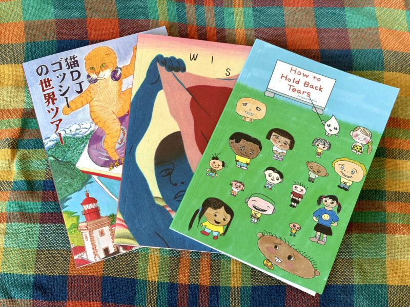 Photo des trois Kuš! Mono disponibles à la micro librairie, l'une est minimaliste, l'autre représente un chat et le dernier des dessins enfantins dans une cour d'école